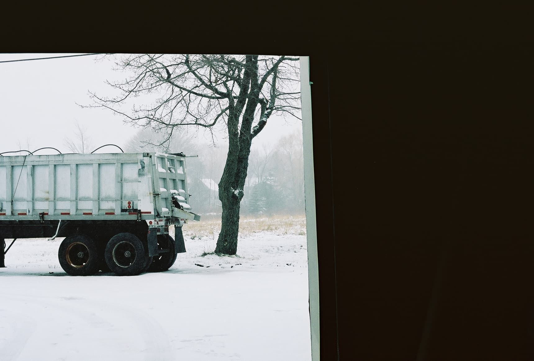 A large industrial truck is parked near a tree in a snow-covered rural area, viewed from inside a dark structure, framing the cold outdoor setting